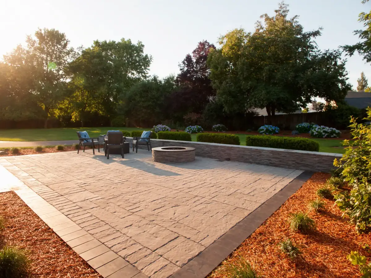 A finished backyard paver patio with a stacked-stone seat wall and gas fire pit at golden hour